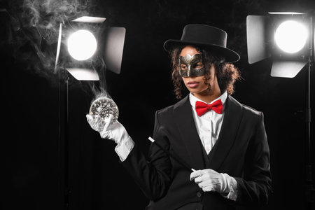 Young African-American magician in mask showing magic trick with crystal ball on black backgroundの写真素材