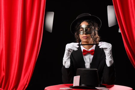 Young African-American magician in mask with magic wand, hat and cards sitting at table on black backgroundの写真素材