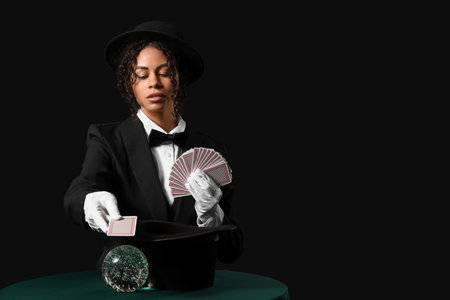 Young African-American magician with cards and crystal ball on table against black backgroundの写真素材