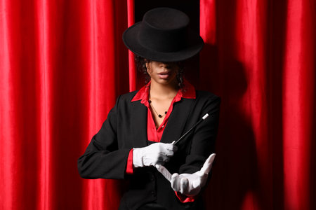 Young African-American magician with magic wand near red curtains on dark backgroundの写真素材