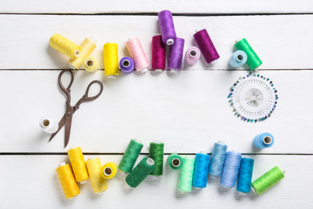 Frame made of colorful thread spools with scissors and sewing pins on white wooden background