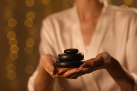 Beautiful young woman with spa stones against blurred garland in dark salon, closeupの写真素材