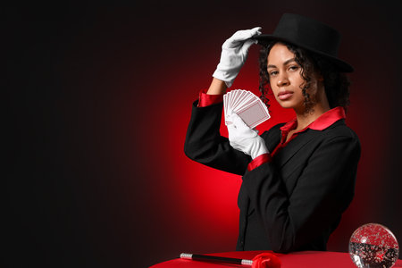 Young African-American magician with magic wand, cards and crystal ball on table against dark red backgroundの写真素材