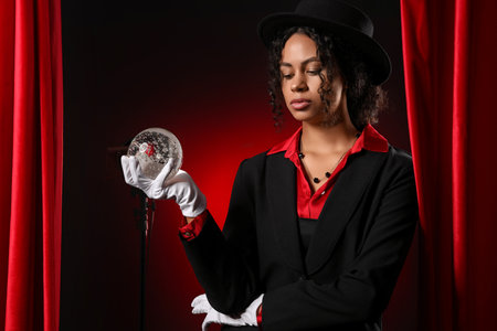 Young African-American magician with crystal ball near red curtains on dark backgroundの写真素材