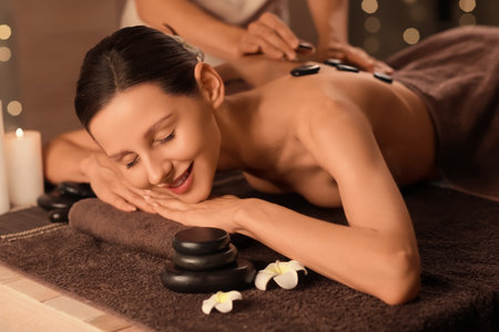 Female masseur giving spa stones massage to woman on couch against blurred garland in dark salonの写真素材