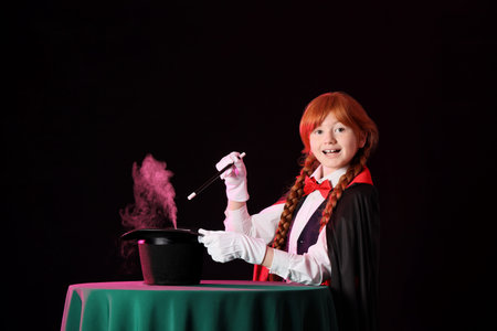 Young magician showing magic trick with hat on table against black background