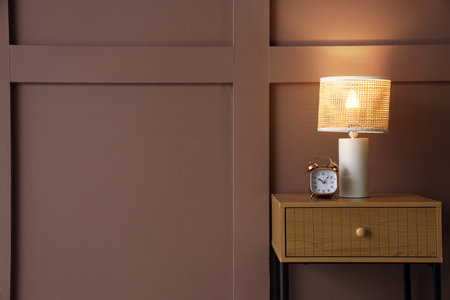 Bedside table with glowing lamp and alarm clock near brown wall