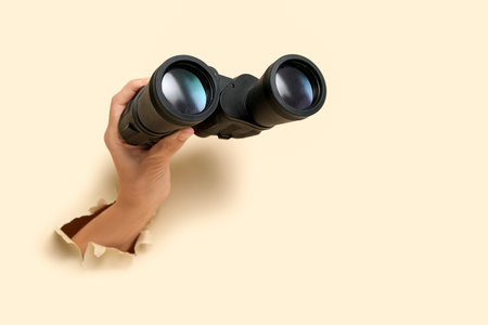 Female hands with modern binoculars visible through hole in beige teared paperの写真素材