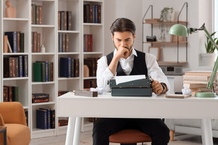 Young male writer with vintage typewriter, books and crumpled papers working in office near bookcase