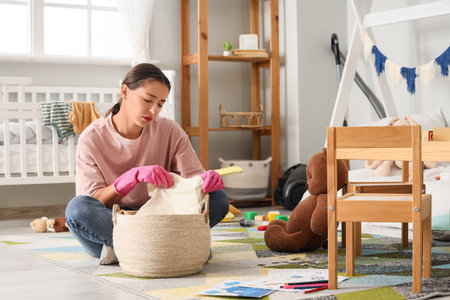 Tired mother with scattered toys and basket of laundry sitting on floor in messy children's roomの写真素材