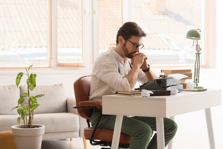 Young male writer with vintage typewriter, crumpled papers and books near window in officeの写真素材