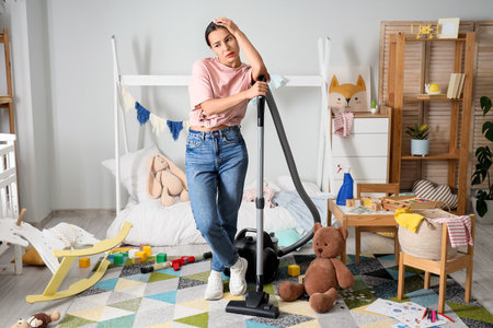 Tired mother with scattered toys, basket of laundry and vacuum cleaner in messy children's roomの写真素材