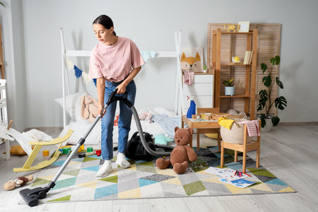 Tired mother with scattered toys and vacuum cleaner cleaning carpet in messy children's roomの写真素材