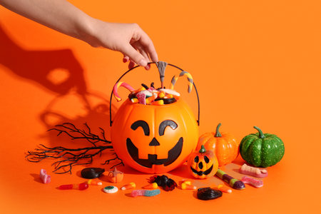Female hand holding cauldron in shape of pumpkin with candies for Halloween on orange backgroundの写真素材
