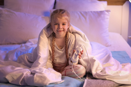 Cute little girl with blanket sitting on electric heating pad in bedroom at nightの写真素材