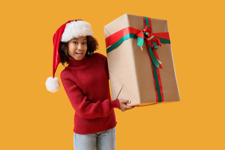 Teenage African-American girl in Santa hat with Christmas gift on yellow backgroundの写真素材