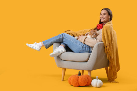Young woman with cute cat in armchair and pumpkin on yellow backgroundの写真素材