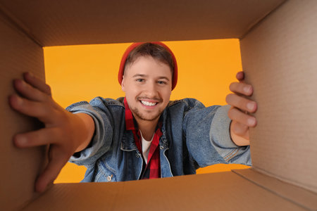 Handsome man with open cardboard box on yellow background, view from inside