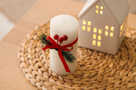 Candle with Christmas branch and house figure on table in room, closeup