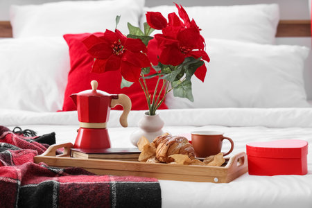 Tasty breakfast with croissant, book and Christmas plant poinsettia on bed in roomの写真素材