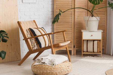 Interior of room with armchair, houseplants, folding screen, wicker ottoman and bedside table near white brick wallの写真素材