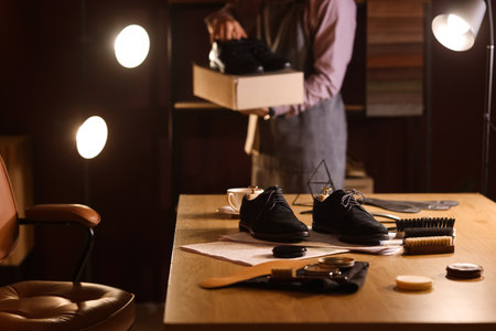 Boots with shoe polishes and brushes on table against male shoemaker in dark workshopの写真素材