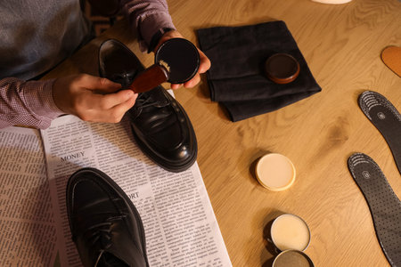 Male shoemaker with brush polishing shoes in dark workshop, top view