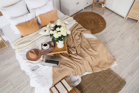 Tray with white roses, coffee, laptop and accessories on bed in room, top view