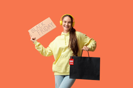 Happy young woman in headphones holding poster with text BLACK FRIDAY and shopping bag on orange backgroundの写真素材