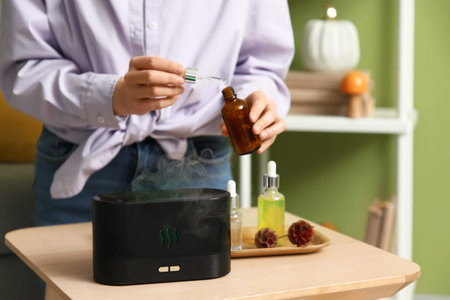 Woman with flowers adding essential oil to air humidifier on table near green wall in room, closeup