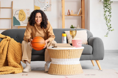 Female African-American sports fan with ball watching basketball on sofa at homeの写真素材