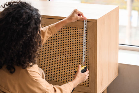 Young African-American woman measuring chest of drawers at home