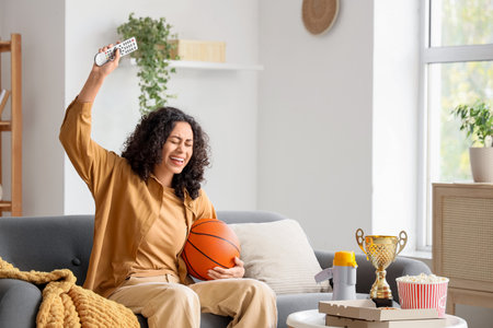 Female African-American sports fan with ball watching basketball on sofa at homeの写真素材
