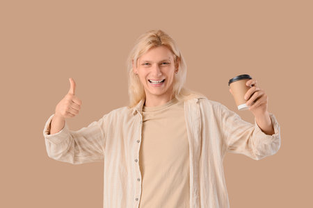Young blonde man with coffee cup showing thumb-up on beige backgroundの写真素材