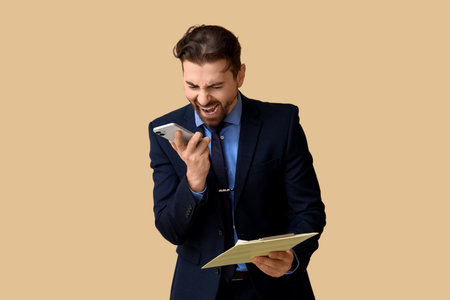 Angry businessman with clipboard shouting at mobile phone on beige backgroundの写真素材