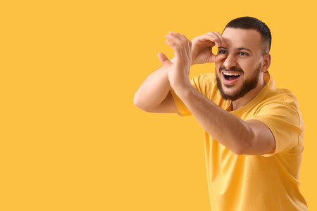 Young man looking through imaginary telescope on yellow backgroundの写真素材