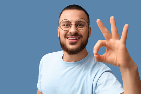 Young man in eyeglasses showing OK on blue background, closeupの写真素材