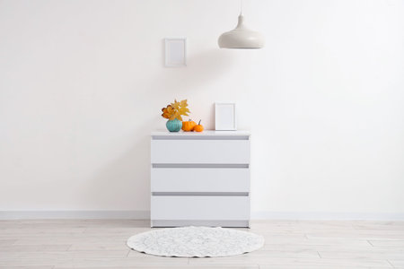 Interior of light room with autumn leaves in vase and pumpkins on dresser