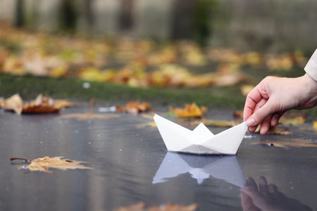 Female hand putting paper boat on puddle with autumn leaves outdoors, closeupの写真素材