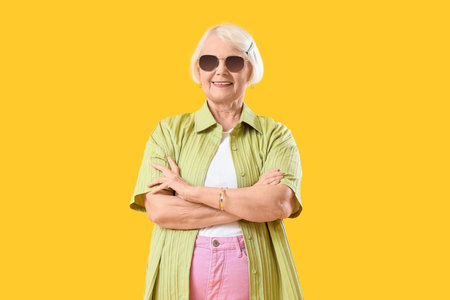 Stylish senior woman in sunglasses on yellow background