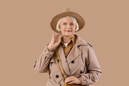 Senior woman in stylish trench coat with hat and bag on beige backgroundの写真素材