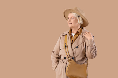 Senior woman in stylish trench coat with hat and bag on beige backgroundの写真素材