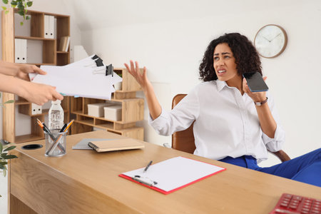 Angry African-American businesswoman with mobile phone displeased of papers at desk in officeの写真素材