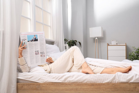 Young woman with newspaper lying in bedroomの写真素材