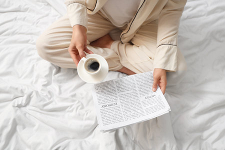 Young woman with cup of coffee and newspaper sitting on bed, top viewの写真素材