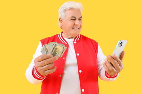 Happy senior man with mobile phone and money on yellow background. Sports bet concept