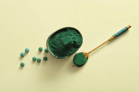 Bowl of spirulina powder with tablets and spoon on green background