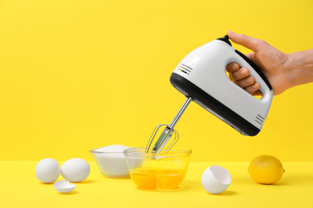 Female hand with modern mixer, bowl of sugar, lemon and eggs on yellow backgroundの写真素材