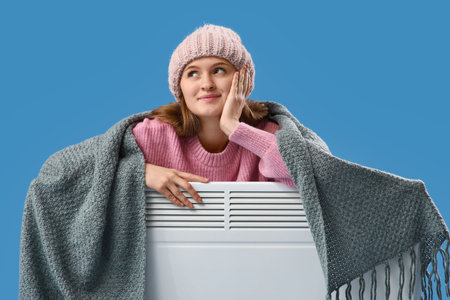Young woman in hat with plaid and electric convector heater on blue background