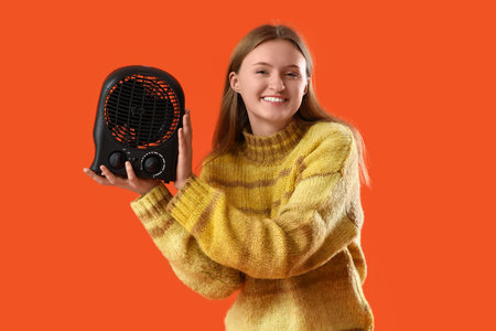 Young woman in sweater with black electric fan heater on orange backgroundの写真素材